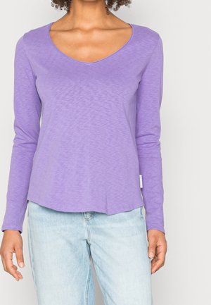 Frau trägt ein langärmliges Lavendel-V-Ausschnitt-Shirt kombiniert mit hellblauen Jeans, stehend vor einem einfarbigen Hintergrund.