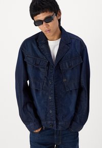 Jachetă din denim închis, cu un guler structurat, două buzunare pe piept și închidere cu nasture. TexturăNetedă și mâneci lungi. Purtați peste o cămașă albă.