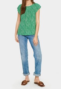 Top verde a maniche corte con un motivo a zigzag, abbinato a jeans azzurri arrotolati e sandali scivolati marroni, che mostrano un outfit informale.