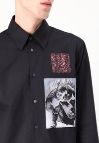Black button-up shirt featuring a textured pink patch and a graphic print of a sculpture. Fabric appears smooth, with a classic collar design.