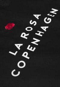 Tessuto nero con testo bianco "LA ROSA COPENHAGEN" e un piccolo dettaglio di rosa ricamata rossa. Il testo è in grassetto e di stile moderno.