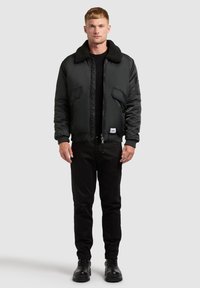 Jachetă bomber neagră cu guler din blană, închidere cu fermoar și buzunare laterale. Purtați-o peste o cămașă neagră, asociată cu pantaloni negri și cizme.