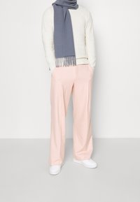 Un pantalon tailleur rose clair à la coupe décontractée, associé à un pull en tricot crème et une écharpe grise à franges. Des baskets blanches complètent le look.