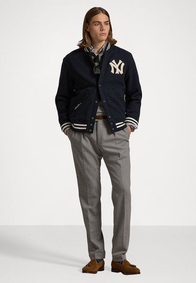 Marineblå uld varsity-jakke med cremefarvede stribede manchetter og New York-logo, parret med grå habitbukser og brune loafers, stående på en ensfarvet baggrund.