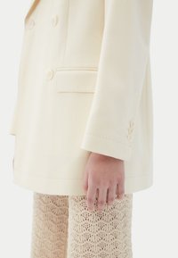 Blazer crème croisé à quatre boutons, avec une poche latérale. Associé à un pantalon beige en maille présentant un motif ondulé.