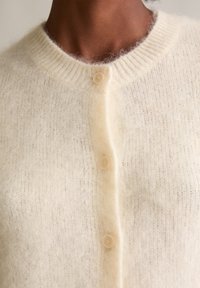 Kardigan w kolorze kremowym, wykonany z miękkiego, puszystego materiału. Posiada ryflowany dekolt i pięć beżowych guzików z przodu.