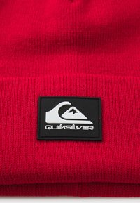 Berretto a maglia rosso con finitura testurizzata, dotato di un'etichetta rettangolare nera con un logo bianco e il nome del marchio "Quiksilver."