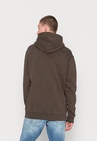 Brauner Hoodie mit Kapuze, gerippten Bündchen und Saum. Das Material wirkt weich mit einer glatten Textur und wird mit hellblauen Jeans kombiniert.