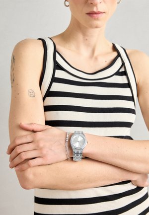 Montre en argent avec un cadran rond et un bracelet en métal maillé, portée avec un bracelet en argent, sur fond d'un haut à rayures noires et blanches.