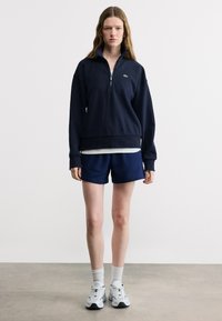 Νεαρή γυναίκα που φοράει μπλε σκούρο μισό φερμουάρ φούτερ, μπλε σκούρα σορτς, λευκές κάλτσες και λευκά αθλητικά παπούτσια, στέκεται μπροστά σε ένα απλό λευκό τοίχο.