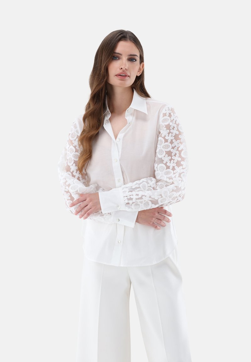 Blusa bianca con maniche in pizzo floreale trasparente, chiusura con bottoni e collo alla francese. Tessuto leggero con una vestibilità su misura.