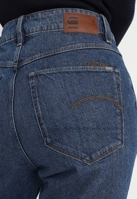 Jeans de mezclilla oscura con cintura alta, con un parche de cuero marrón que dice "RAW", costuras visibles y un solo bolsillo trasero.