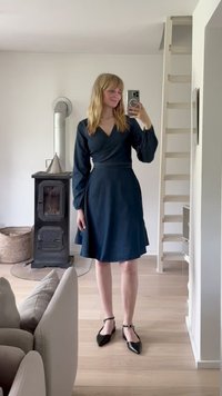 Marineblå omslagskjole med lange ermer og en utsvingt skjørt, har et teksturert stoff. Modell iført svarte spisse flats står i et lyst rom.