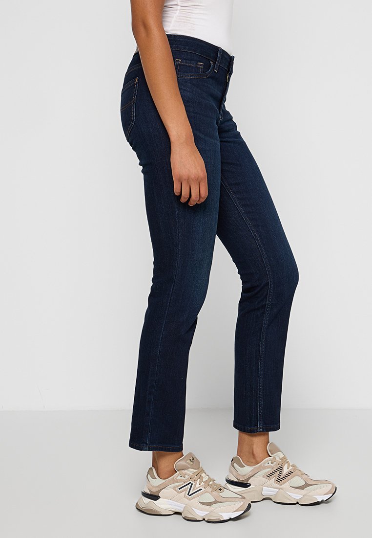 Jeans skinny in denim blu scuro, con vita a media altezza, dettagli di cucitura sottili e tasche laterali. Abbinati a sneakers beige.