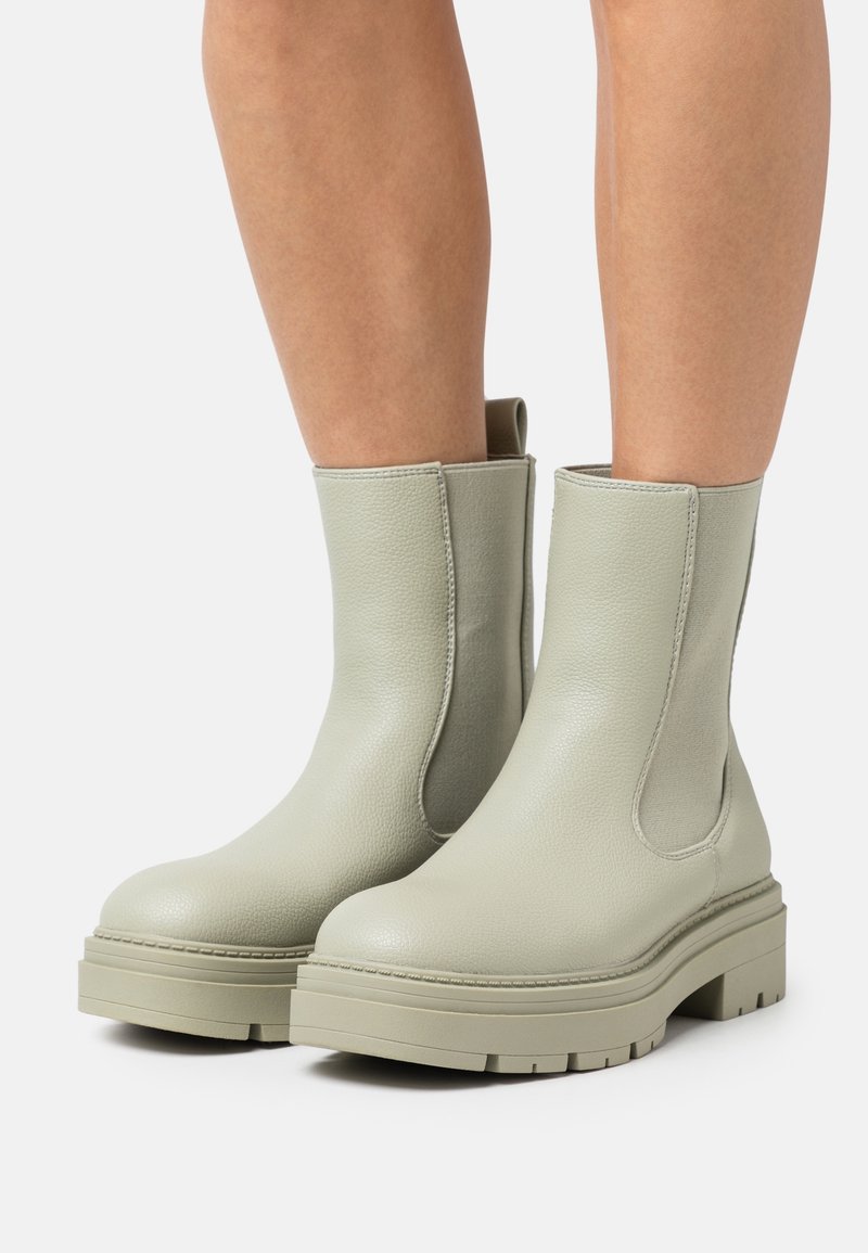 Bottines en cuir gris clair avec semelles épaisses portées sur jambes nues, présentées sur un fond clair uni.