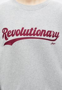 Γκρι φούτερ με τη λέξη "Revolutionary" κεντημένη με μεγάλα κόκκινα γράμματα σε πλάγια γραφή στο στήθος.