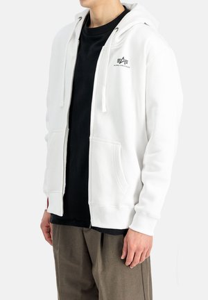 Person trägt einen weißen Kapuzenpullover mit Reißverschluss und Fronttaschen sowie ein schwarzes Shirt darunter, braune Hose, steht vor einem weißen Hintergrund.