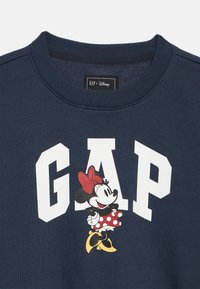 Tumši zils džemperis ar rāvējslēdzēja kaklu, uz kura ir liels balts uzraksts "GAP" un grafiskais attēls ar Mini Mūsu sarkanā polka punktu kleitā.