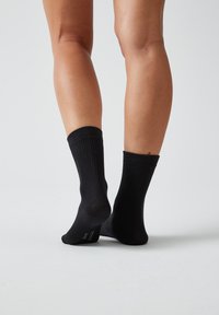 Calze nere in cotone con texture a coste e altezza alla caviglia, caratterizzate da una soletta liscia, indossate a piedi nudi su uno sfondo chiaro.