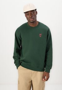 Green sweatshirt with long sleeves, ribbed cuffs, and a crew neck. Features a small red embroidered design on the chest. Casual fit.