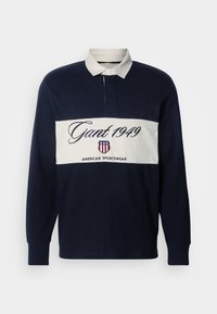 Polo cu mânecă lungă de culoare bleumarin, cu guler alb și bandă orizontală albă. Prezintă logo-ul "Gant 1949" și brandingul American Sportswear. Material dintr-un amestec de bumbac.