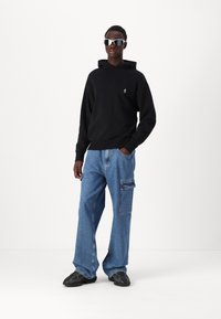 Sweatshirt à capuche noir, jeans cargo bleu clair à jambes larges avec poche latérale, et baskets noires. Le modèle porte des lunettes de soleil argentées.