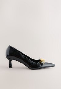 Escarpin mi-hauteur à bout pointu en cuir verni noir avec un détail chaîne décorative dorée à l'avant.
