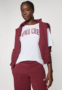 Conjunto de activewear en color burdeos que incluye un hoodie sin mangas sobre una camiseta blanca con el estampado "PUMA CREW" y pantalones de chándal burdeos a juego.