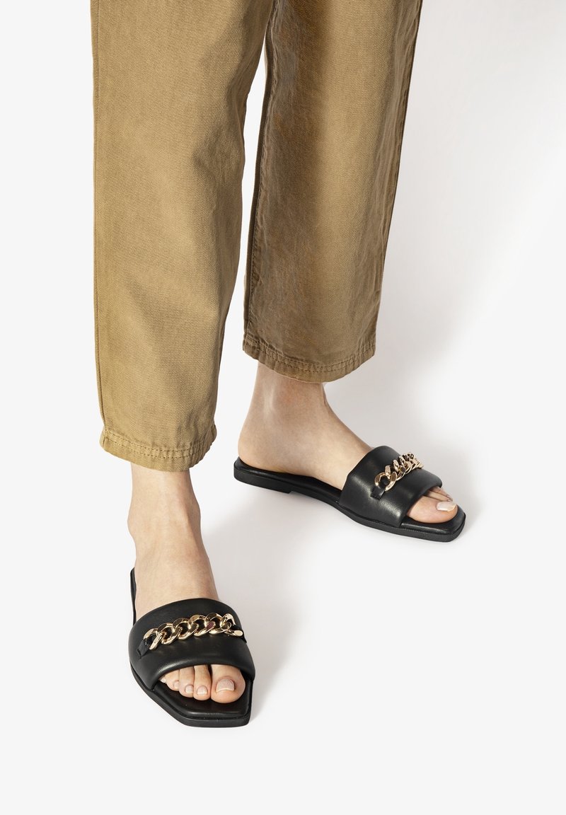 Pieds portant des sandales noires à bout ouvert avec un détail de chaîne dorée, associées à un pantalon marron court sur fond blanc.