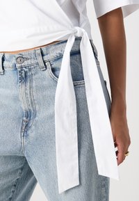 Gros plan sur une personne portant un jean bleu clair et une chemise blanche nouée à la taille, montrant une main avec une bague en or reposant sur le côté.