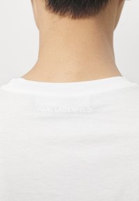 Balta medvilninė T marškinėliai su apvalia iškirpte ir matomu „KARL LAGERFELD“ logotipu iškilmingos baltos spalvos tekstu nugaros pusėje. Sklandi tekstūra.