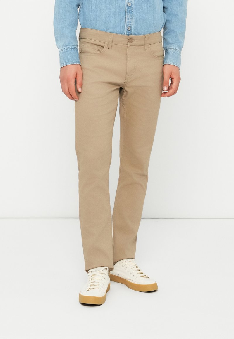 Beige bomullsbukser med slim passform, med knappelukking og frontlommer, kombinert med hvite sneakers med beige såler.