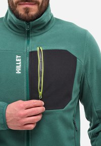 Veste en polaire teal et noire avec un col haut, dotée d'une poche zippée sur la poitrine avec une tirette verte et des textures contrastées.