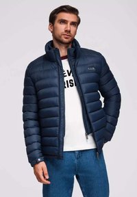 Gepolsterte Jacke in Dunkelblau, mit hochgestelltem Kragen, Reißverschluss und Seitentaschen. Über einem weißen Hemd mit Text getragen. Leichter, glatter Stoff.