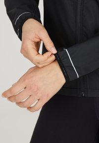 Schwarze Sportjacke mit Reißverschluss auf der Vorderseite, ausgestattet mit elastischen Bündchen mit weißen Akzenten und einer glatten Textur. Hand, die das Bündchen anpasst.