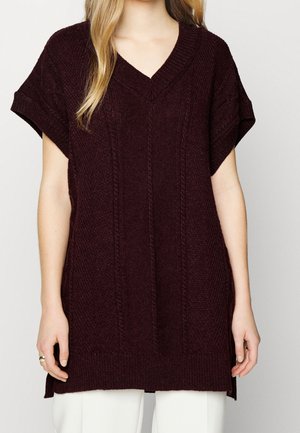 Femme portant un pull sans manches en maille bordeaux foncé à col en V, avec des détails en maille torsadée et texturée, sur un pantalon blanc.