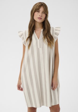 Femme aux longs cheveux bruns portant une robe sans manches beige et blanche à rayures verticales avec des épaules à volants sur un fond uni.