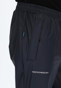 Zwarte waterdichte broek met een elastische tailleband, voorzien van een ritszak en een logo in het wit gedrukt op het been.