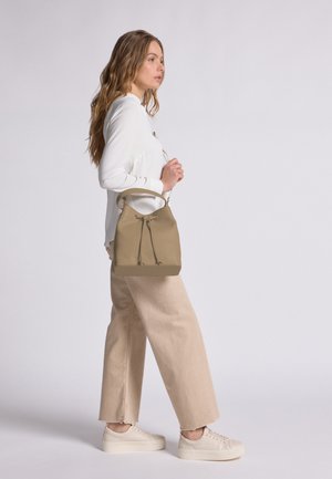 Beige Leder-Einkaufstasche mit Kordelzugverschluss und einem Griff, präsentiert von einem Modell in einem weißen Hemd und beigen Hosen, das seitlich steht.