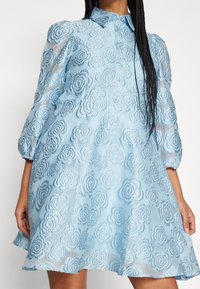 Ljusblå klänning med en texturerad blommönster, knappfront, krage och puffärmar, samt en utsvängd kjol.