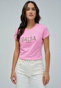 Camiseta de algodón rosa con mangas cortas que presenta "SALSA JEANS" en letras metálicas. Combinada con unos pantalones vaqueros blancos de cintura alta.