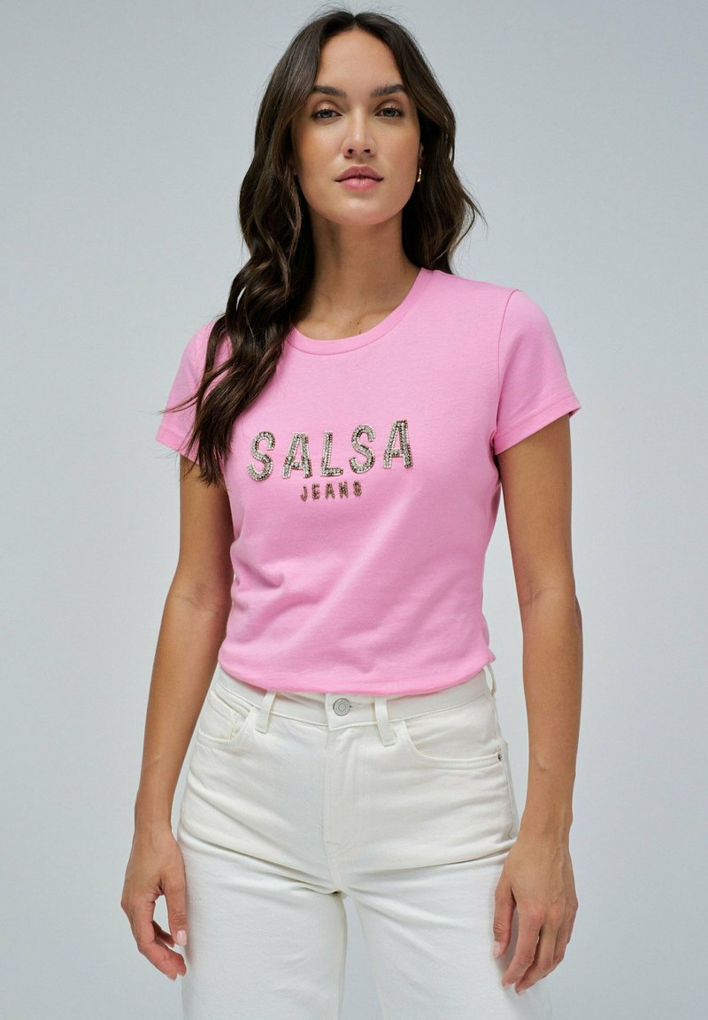 Camiseta de algodón rosa con mangas cortas que presenta "SALSA JEANS" en letras metálicas. Combinada con unos pantalones vaqueros blancos de cintura alta.