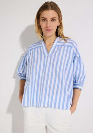 Frau trägt eine lockere, hellblaue und weiße Bluse mit vertikalen Streifen, 3/4-puffärmel und weiße Hose, steht vor einfachem hellem Hintergrund.