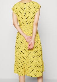 Keltainen pallokuosisella mekolla, jossa on pyöreä pääntie, lyhyet hihat ja nappiyksityiskohta selässä. Kangas on sileää ja malli on A-linjainen.