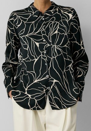 Czarna koszula zapinana na guziki z abstrakcyjnym beżowym wzorem liści, noszona z białymi spodniami, ręce w kieszeniach, na jednolitym szarym tle.