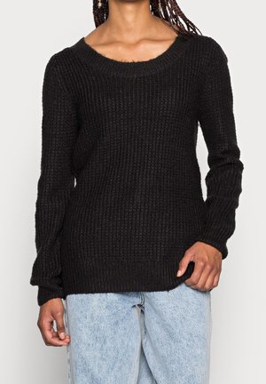 Personne portant un pull en maille côtelée noir à manches longues et un jean bleu clair, debout devant un fond uni.