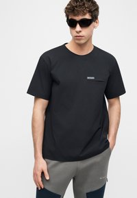 Νέος άνδρας που φοράει μαύρο t-shirt Columbia με τσέπη στο στήθος και γκρι παντελόνι Columbia, ποζάροντας με το ένα χέρι στην τσέπη και φορώντας σκούρα γυαλιά ηλίου.