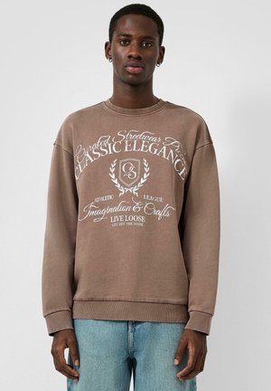 Jonge man die een bruine sweatshirt draagt met witte tekst "Classic Elegance" en een casual blauwe spijkerbroek, staand tegen een eenvoudige lichte achtergrond.