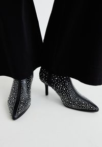 Zwarte enkellaarsjes met een spitse neus, voorzien van zilveren studs versieringen over de hele laars en een hoge stiletto hak. Ritsluiting aan de achterkant.