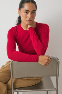 Rotes Langarmshirt mit rundem Ausschnitt, aus glattem Stoff, kombiniert mit beigen Hosen, sitzt auf einem Metallstuhl.